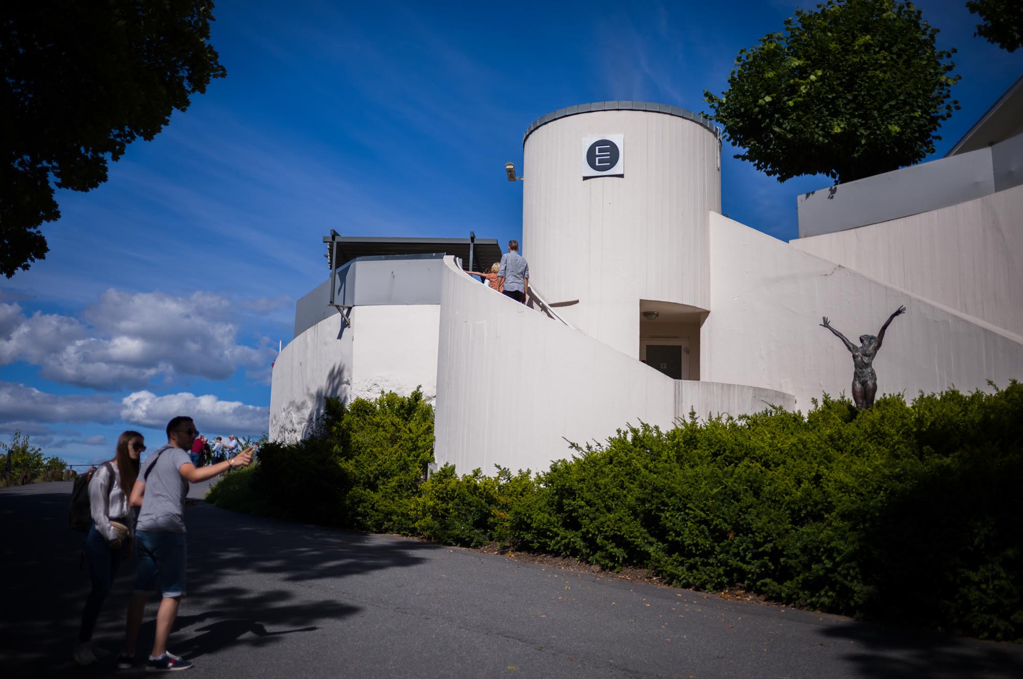 Modern white building with cylindrical design, sculpture in front, and visitors walking on a sunny day.