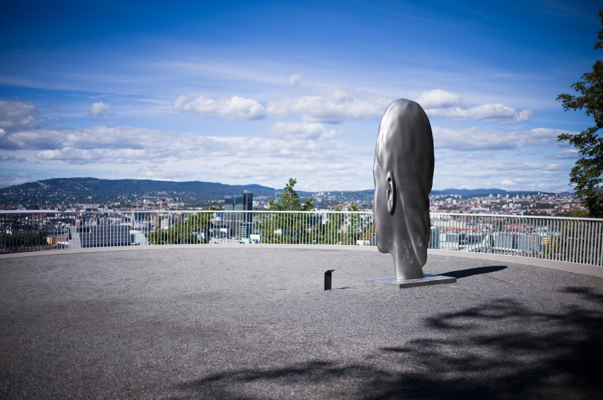 Silver head sculpture on a viewing deck overlooking a cityscape with mountains under a blue sky.