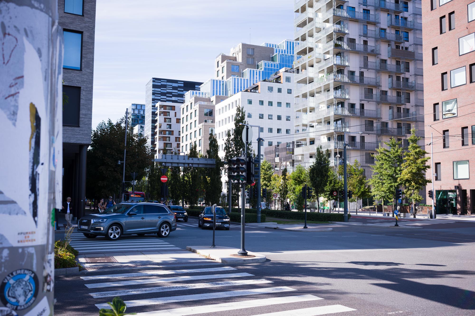 Cityscape with modern apartments, cars at a crosswalk, and trees lining the street under a clear sky.
