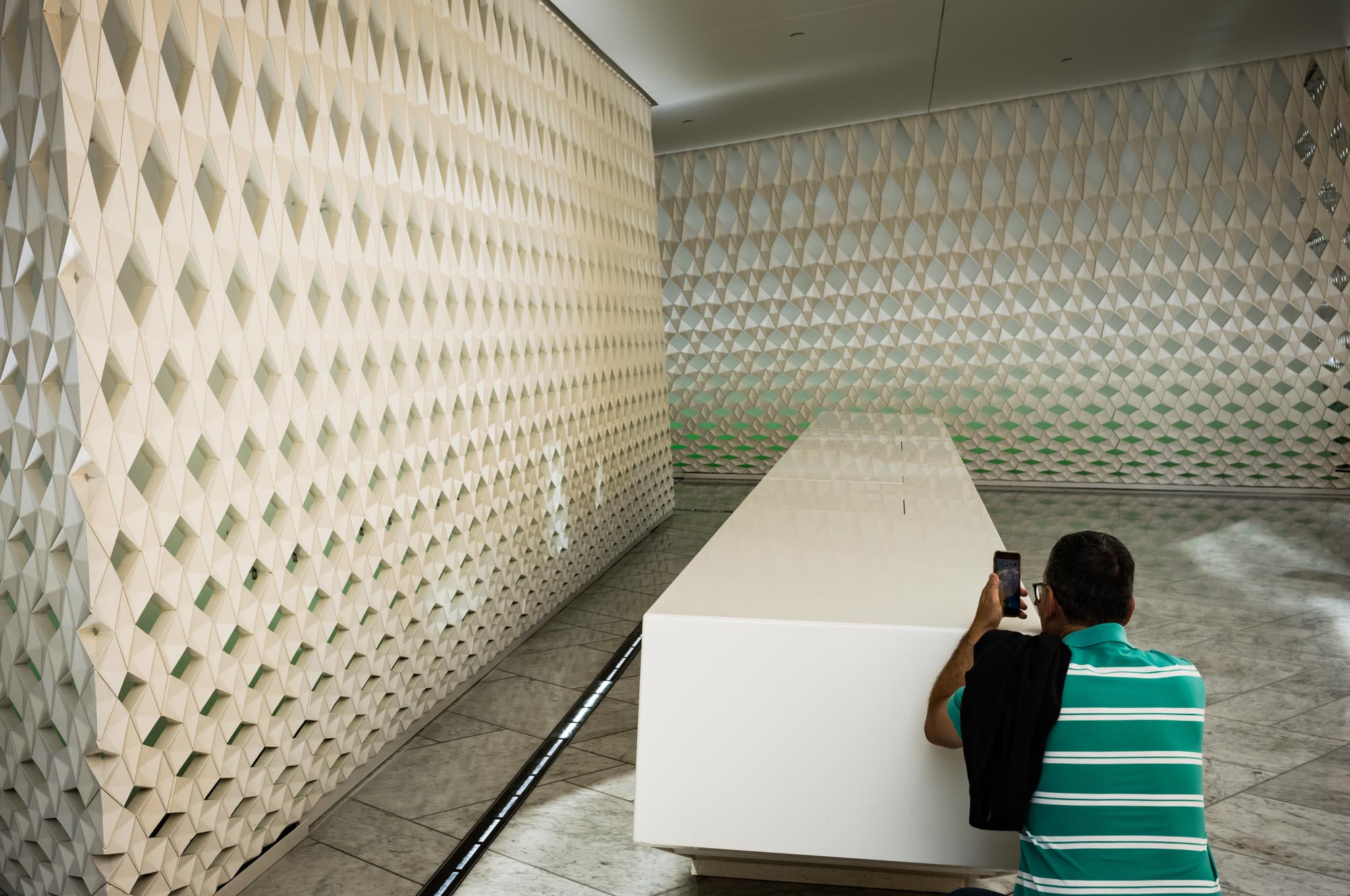 Man in striped shirt taking photo of modern, geometric wall pattern inside building.