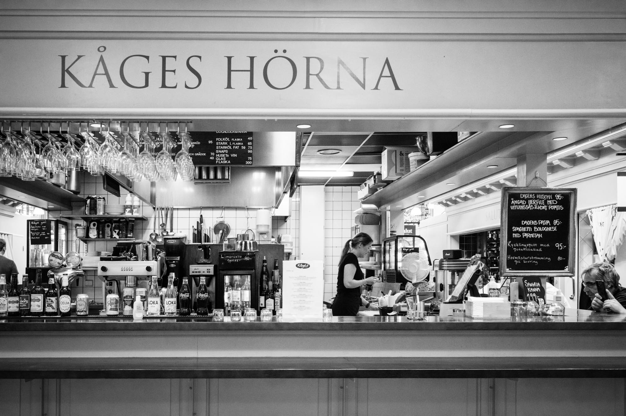Black and white photo of a café counter with drinks and a staff member behind, under the sign 'Kåges Hörna'.