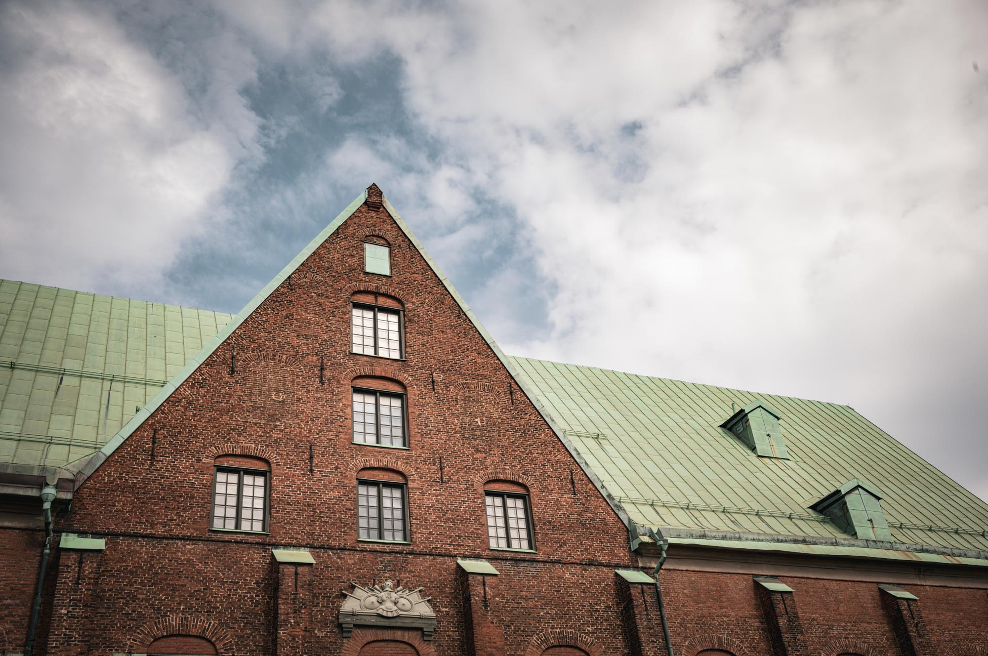 Red brick building with green gabled roof and arched windows under a cloudy sky.