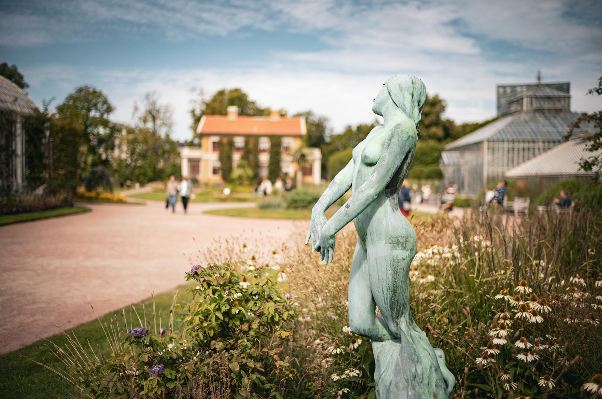 Bronze statue in a garden, surrounded by flowers, with a historic building and greenhouse in the background.