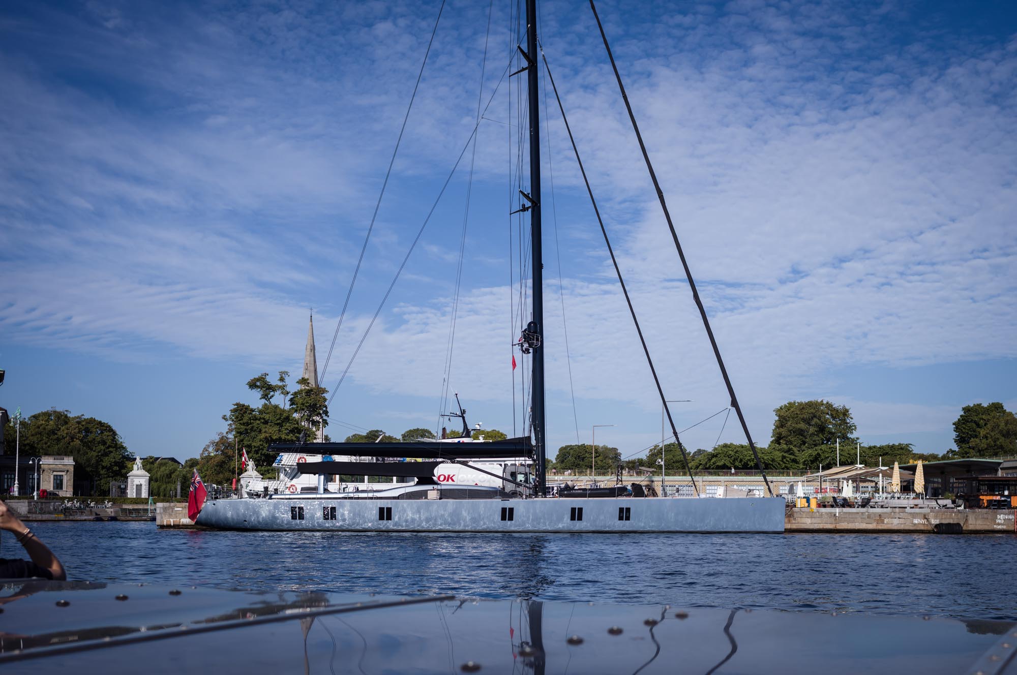 Sleek sailboat docked near a pier with trees and spired building under a blue sky.
