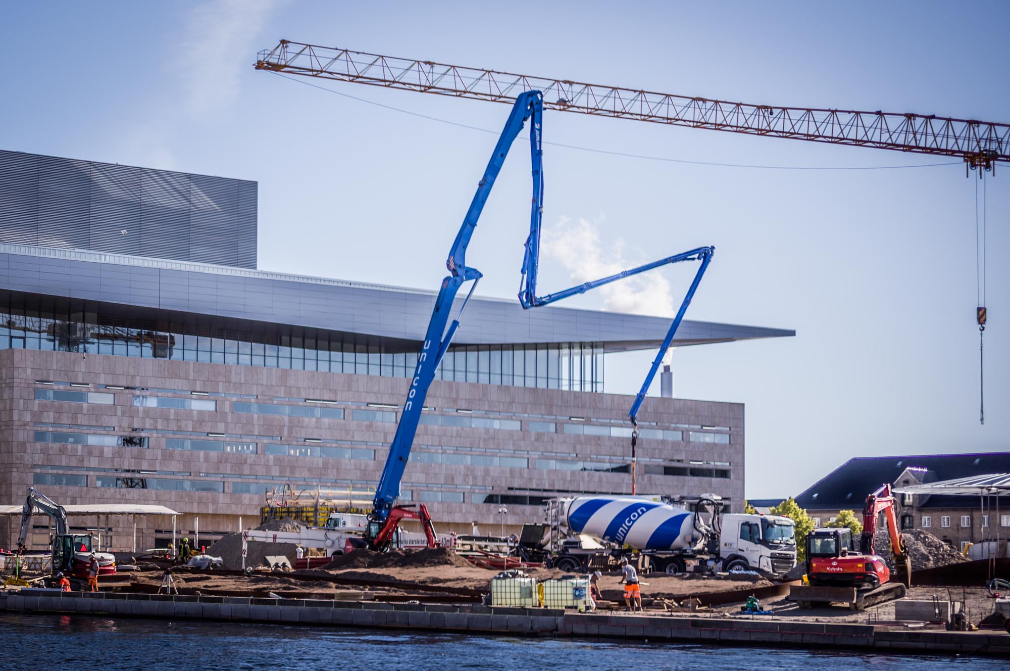 Construction site with cranes and cement mixer near a modern building by waterfront.