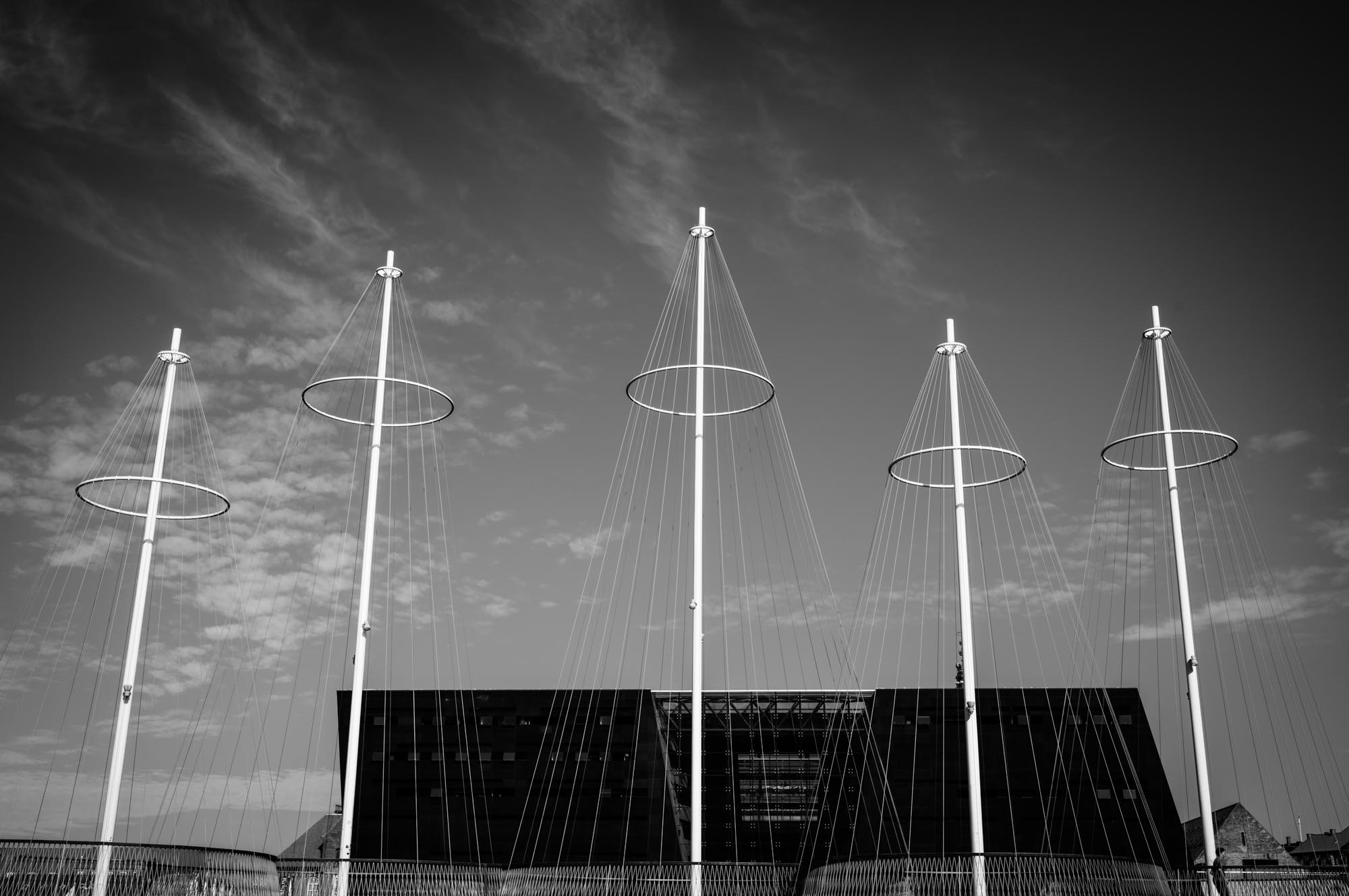 Five modern, tall sculptures with cables and rings against a clear sky, black building in background. Black and white photo.