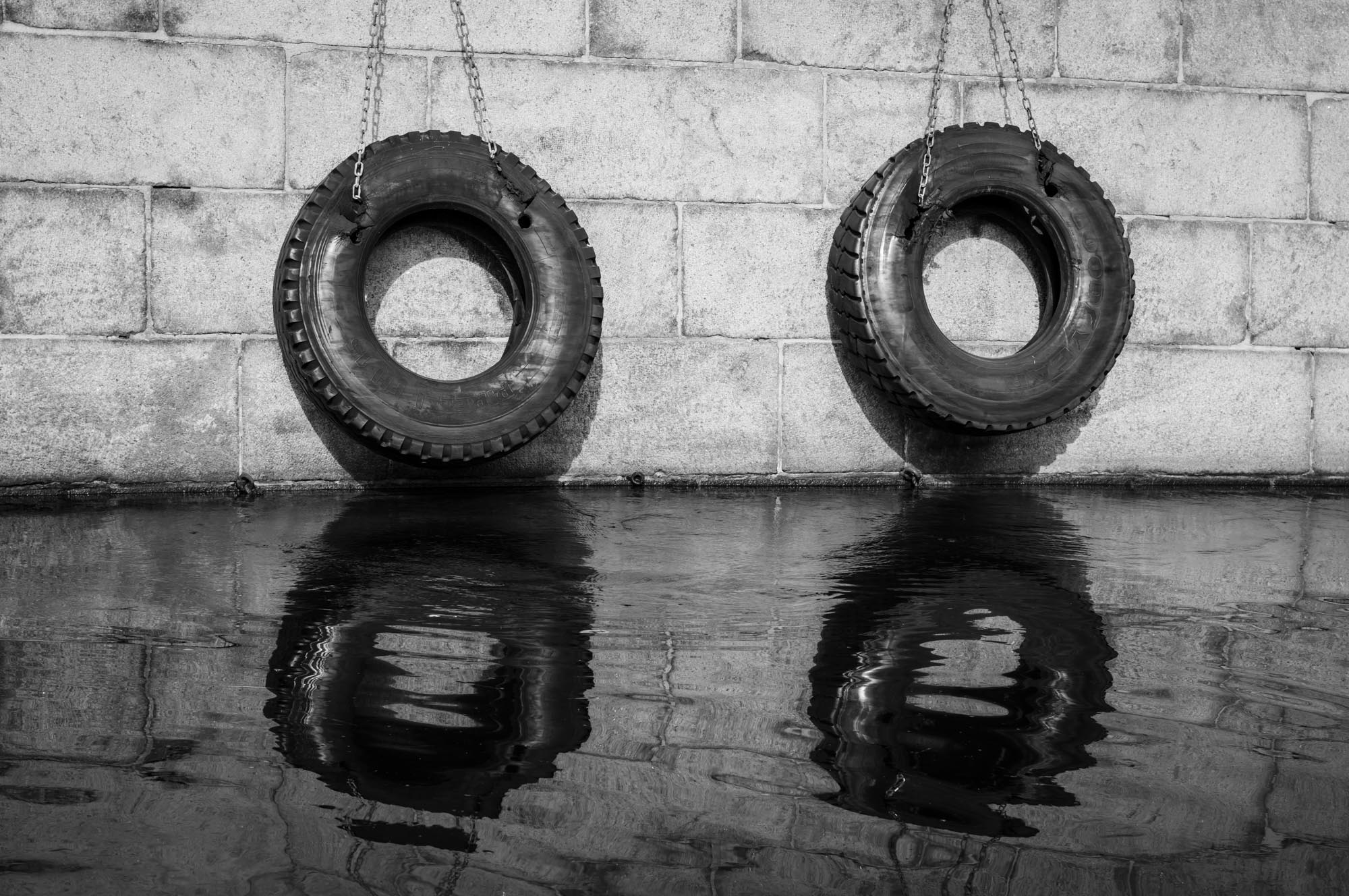 Two hanging tires on chains against a stone wall reflect in calm water. Black and white photography.