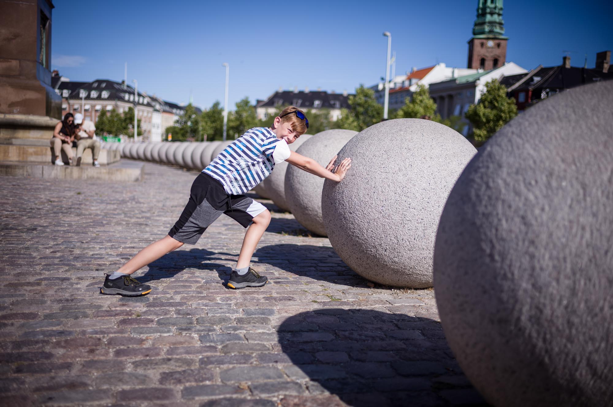 Child in striped shirt playfully pushes large stone ball on cobbled street under blue sky.