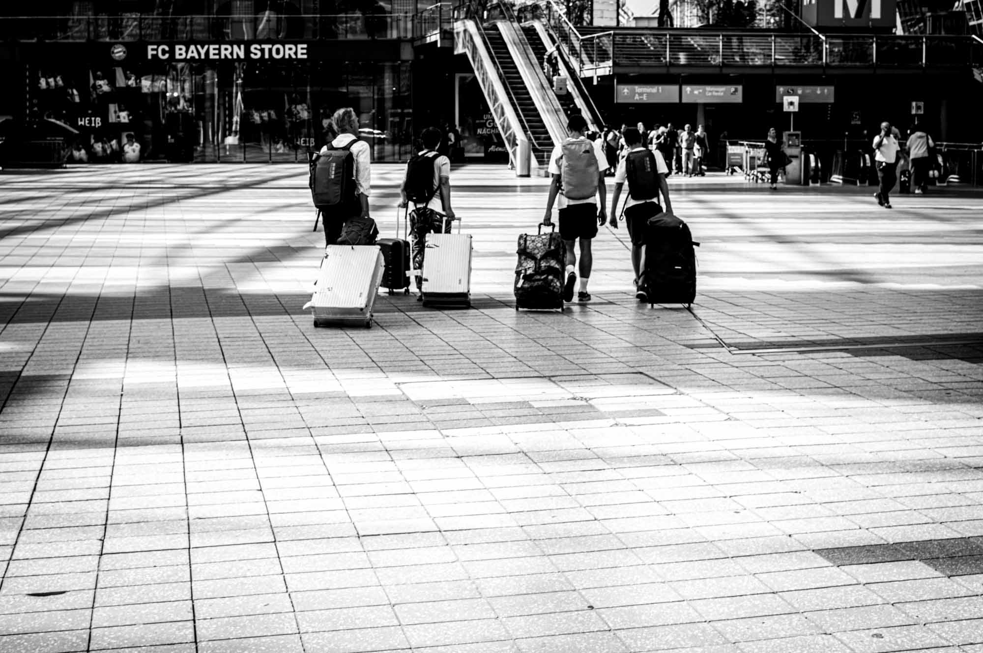 Travelers with luggage in a city square near FC Bayern Store, monochrome scene capturing urban journey.