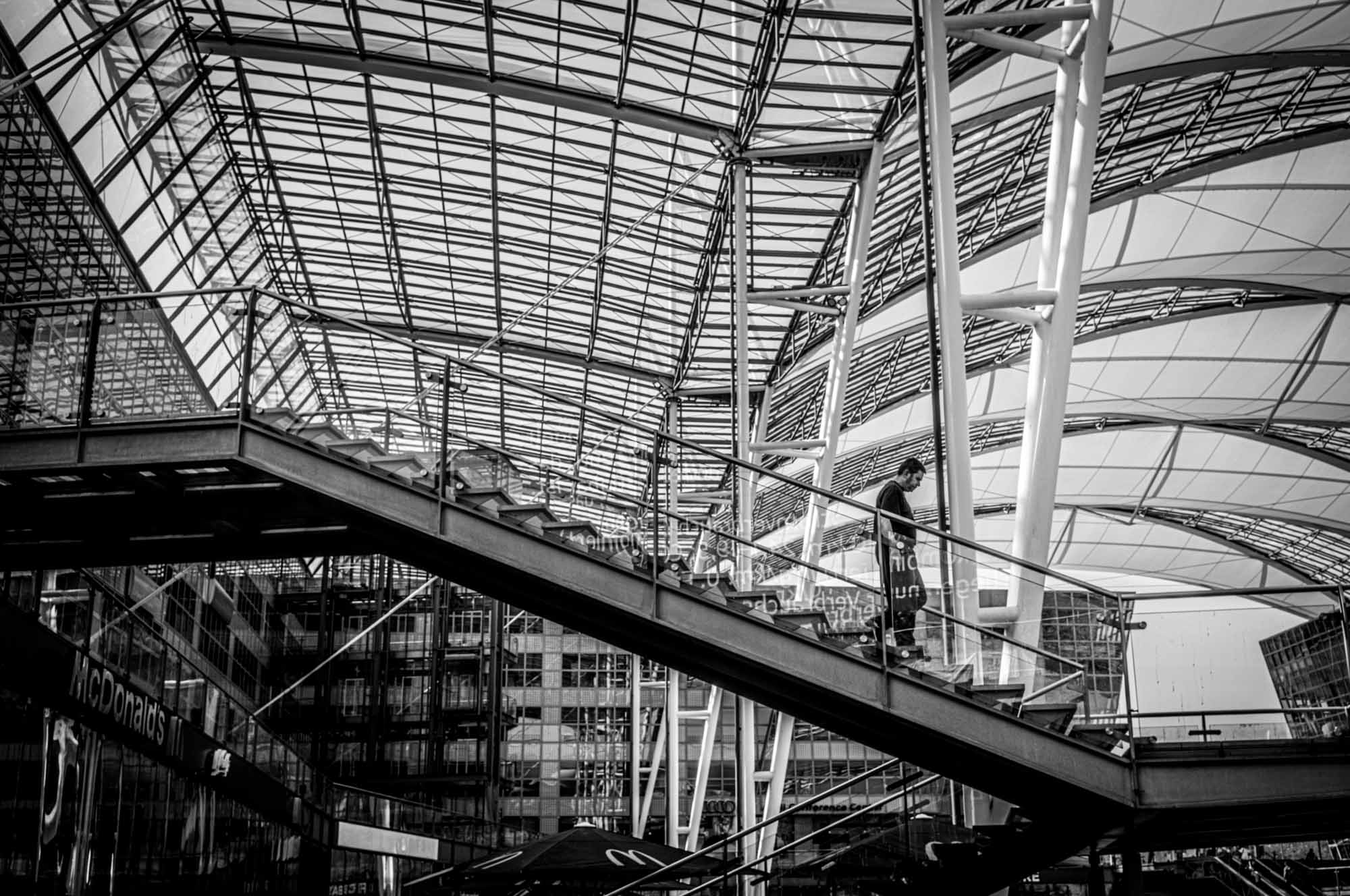 Modern architectural interior with a person on a glass staircase beneath a large, geometric ceiling structure. Black and white photo.