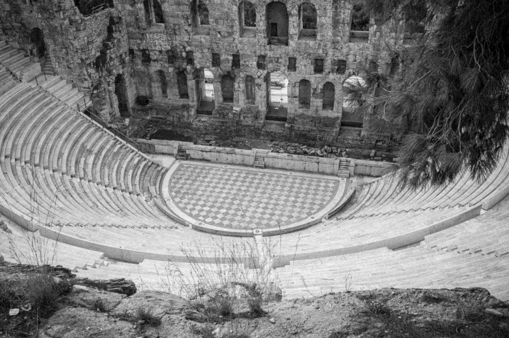 Ancient stone amphitheater with checkerboard-patterned stage and tiered seating, surrounded by ruins and trees.