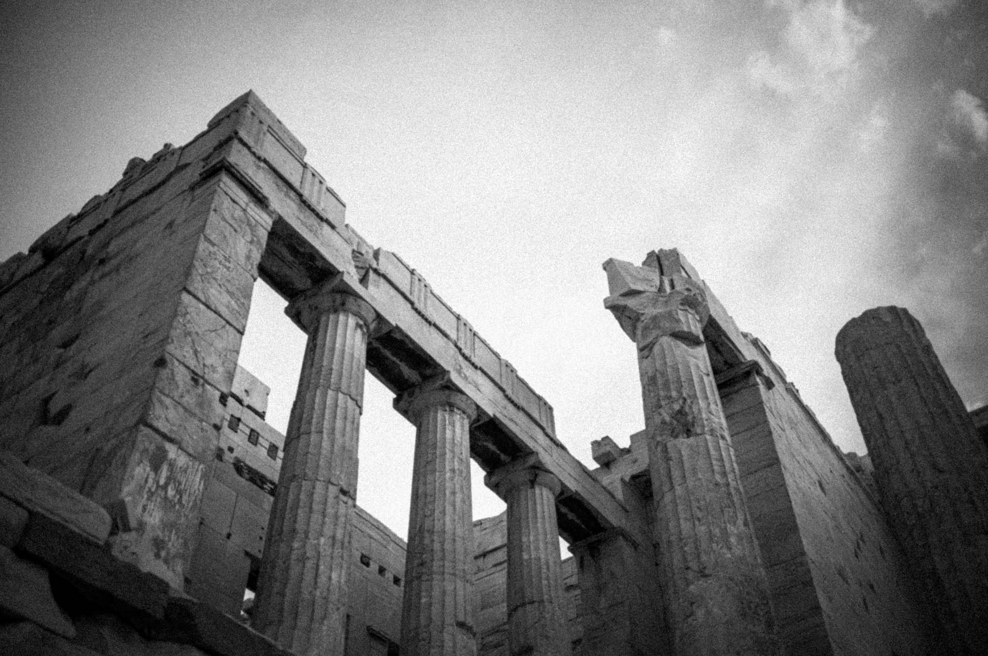 Black and white view of ancient Greek temple ruins with towering columns under a cloudy sky.