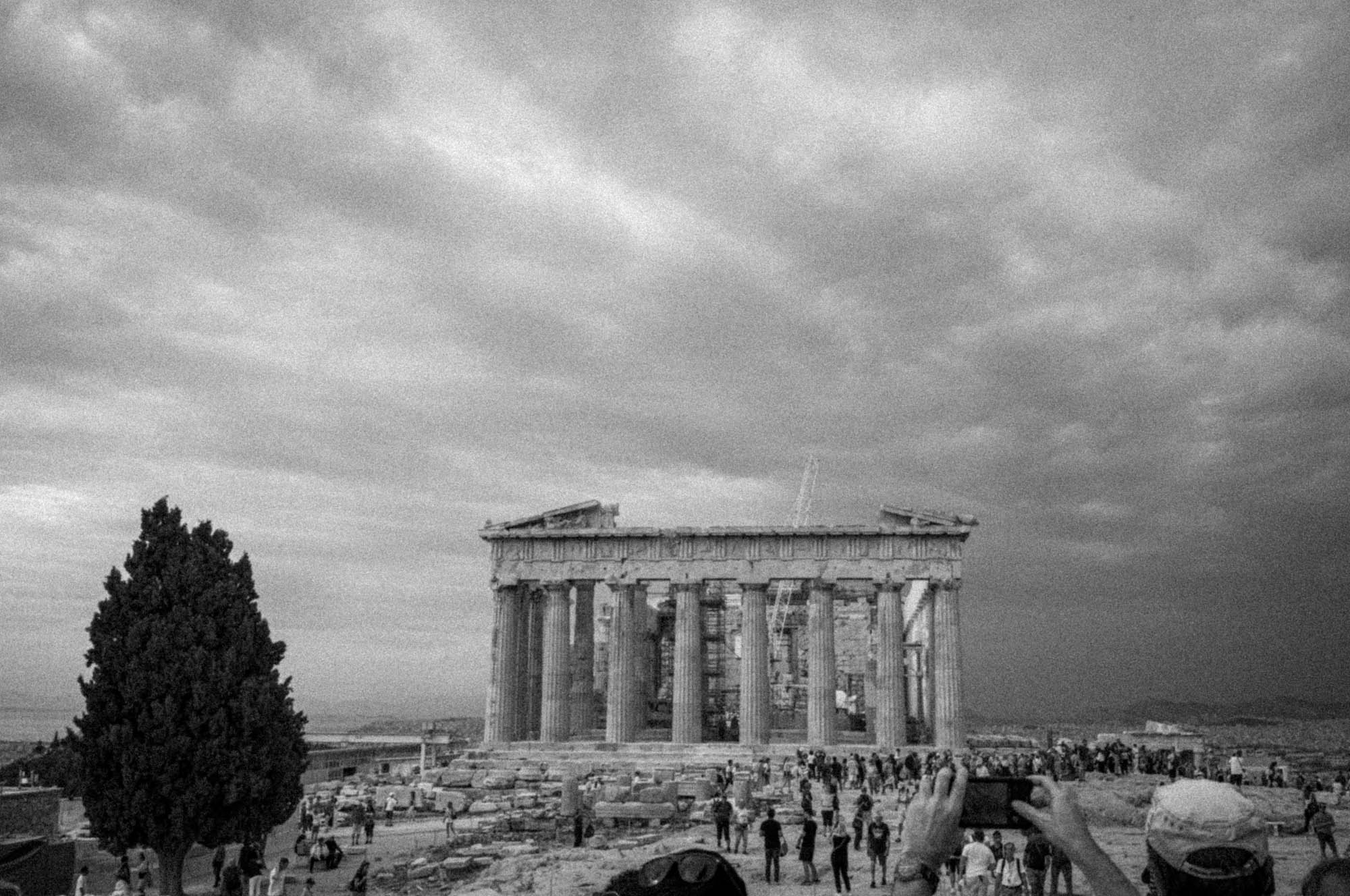Black and white photo of the Parthenon in Athens, with tourists gathered and a dramatic cloudy sky overhead.