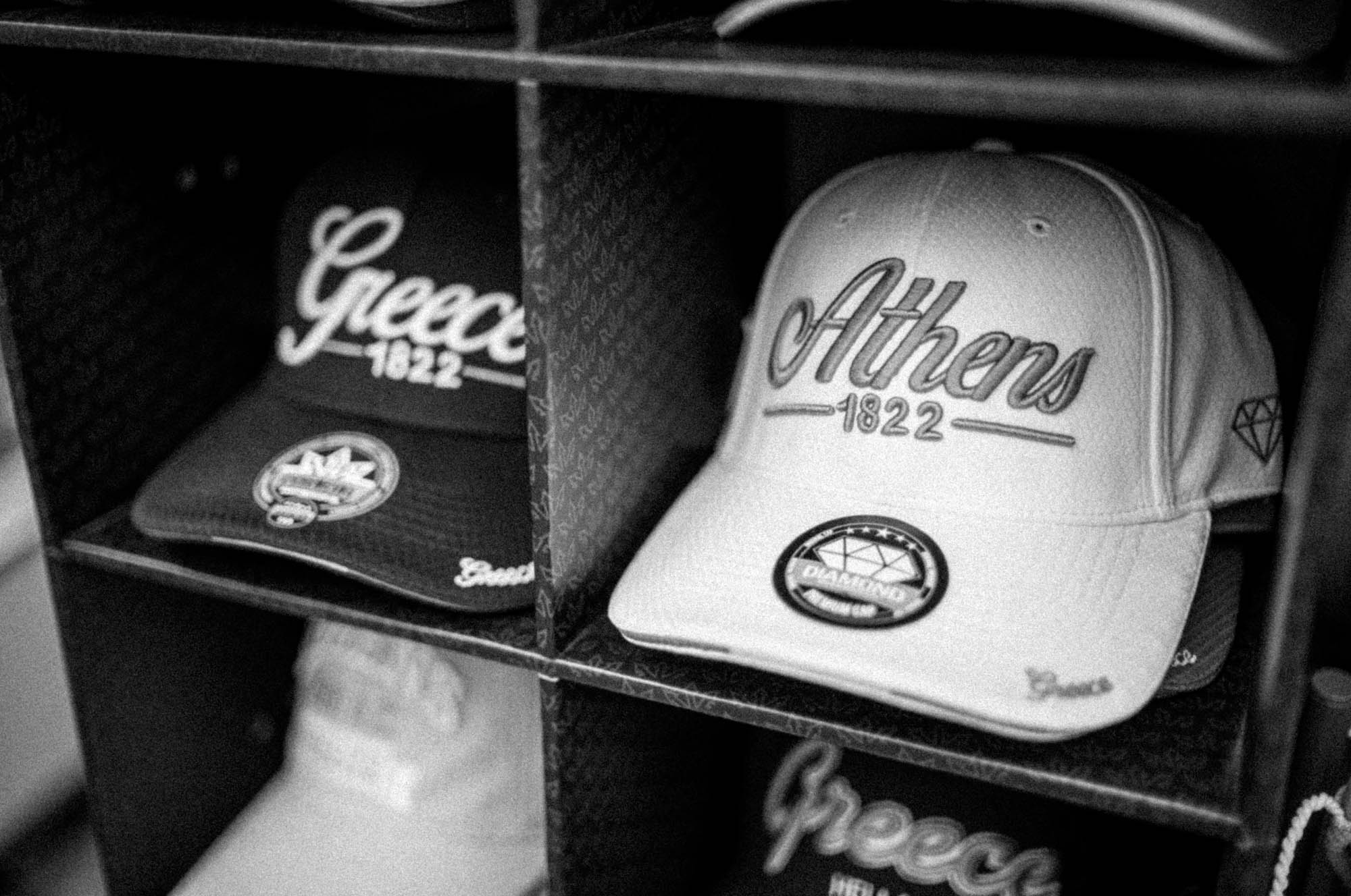 Baseball caps on shelves, “Athens 1822” and “Greece 1822” designs, monochrome image.