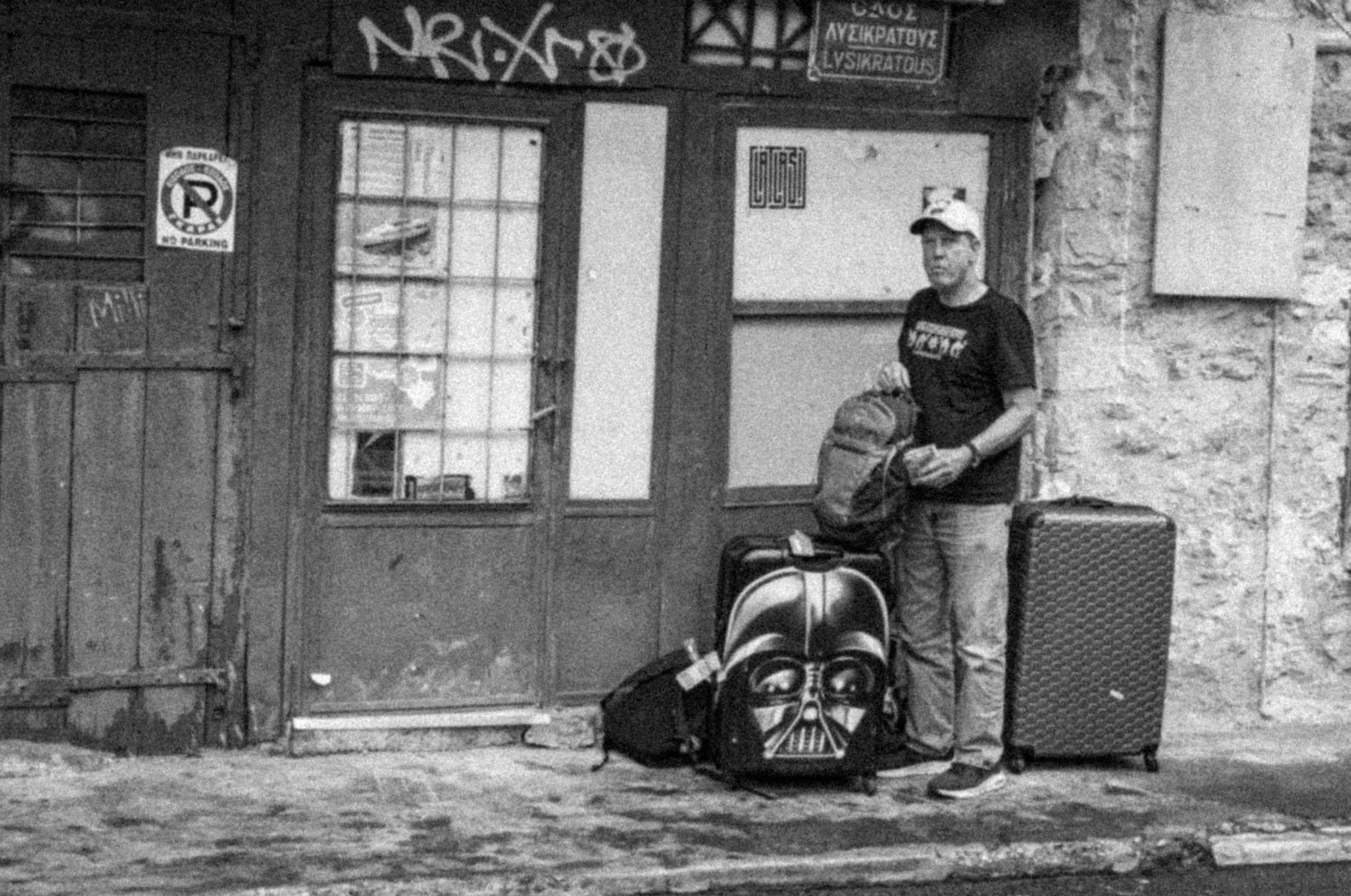Person with Darth Vader luggage and backpack stands in front of an old building, city street scene.