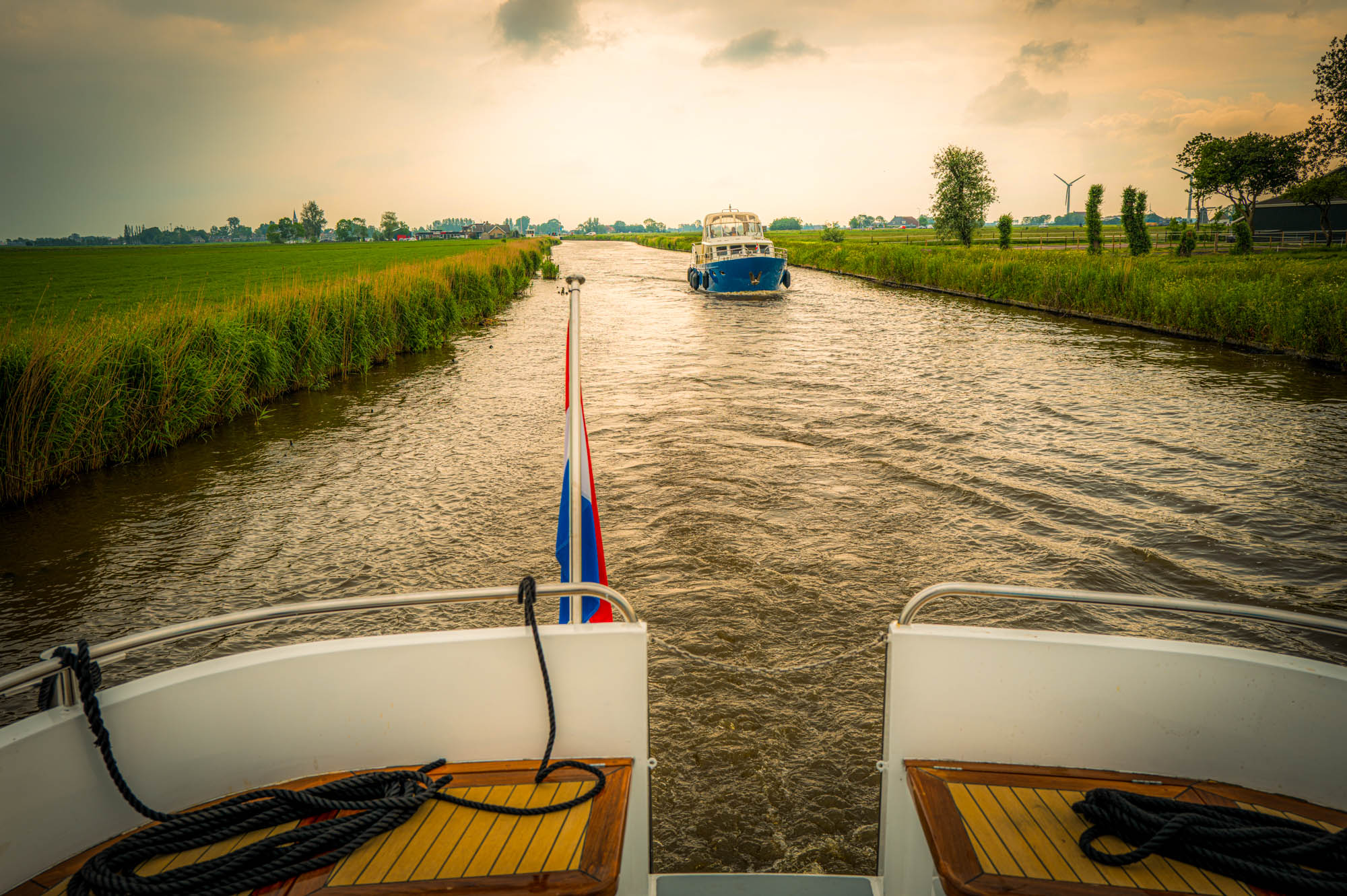Boat cruising a scenic canal in the Netherlands with fields and trees at sunset.