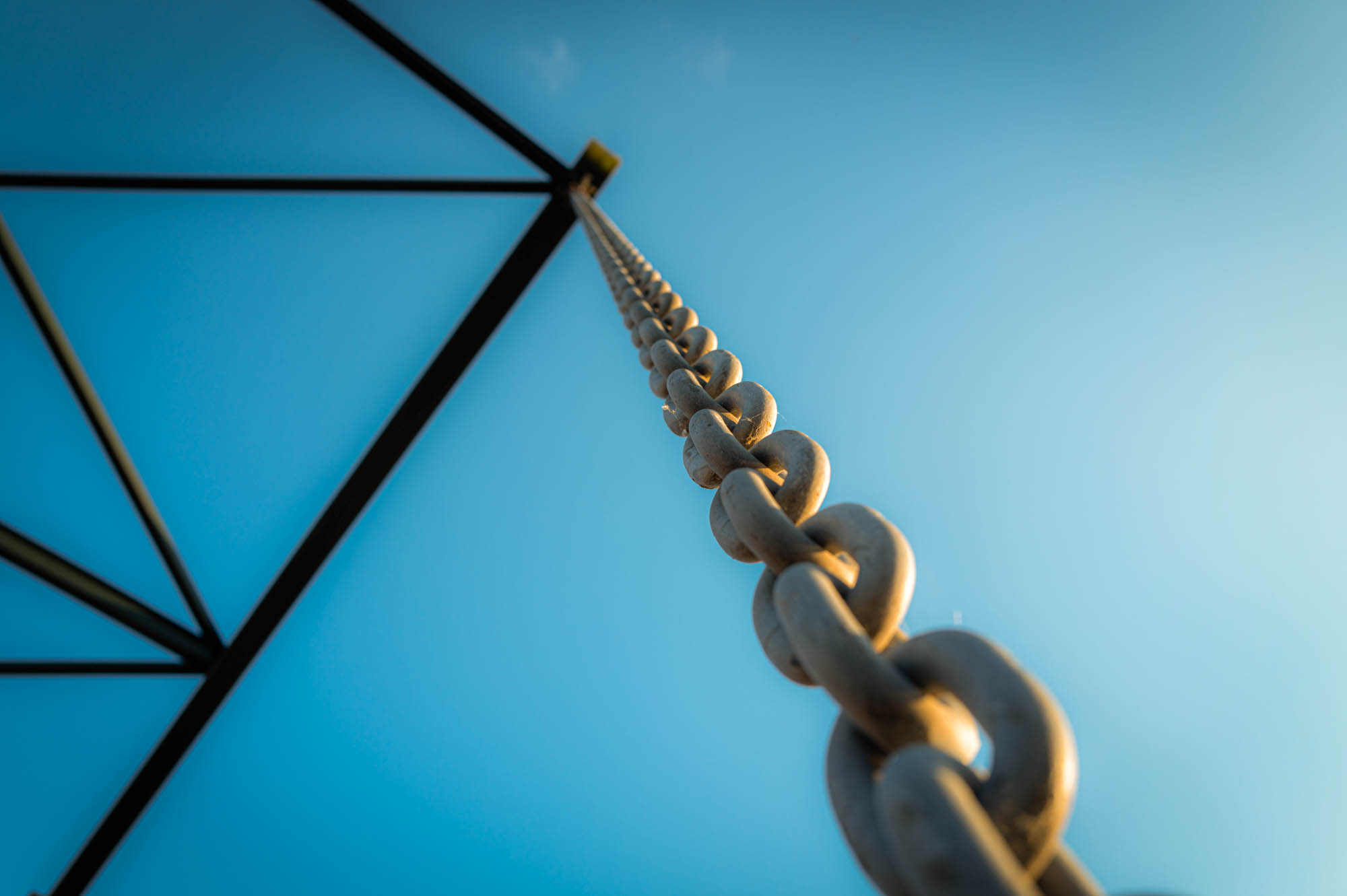 View of a long metal chain extending upward against a clear blue sky background; focus on strength and length.