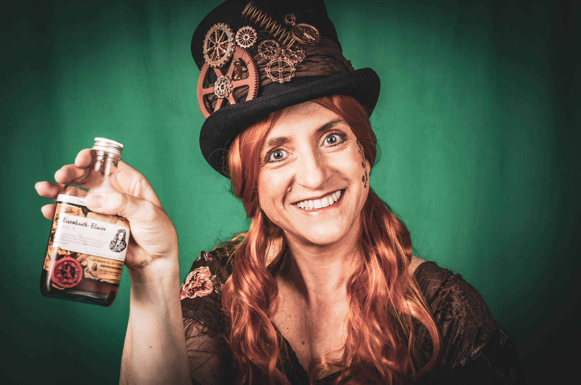 Woman in steampunk attire with decorative hat, holding a vintage bottle against a green background, smiling broadly.