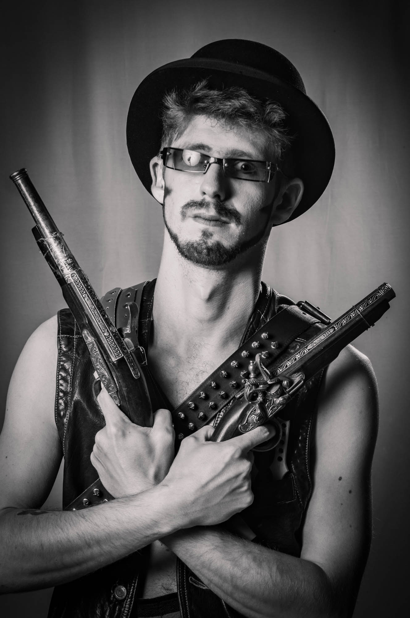 Steampunk-themed man with a hat and glasses holding vintage pistols in a dramatic, black and white portrait.