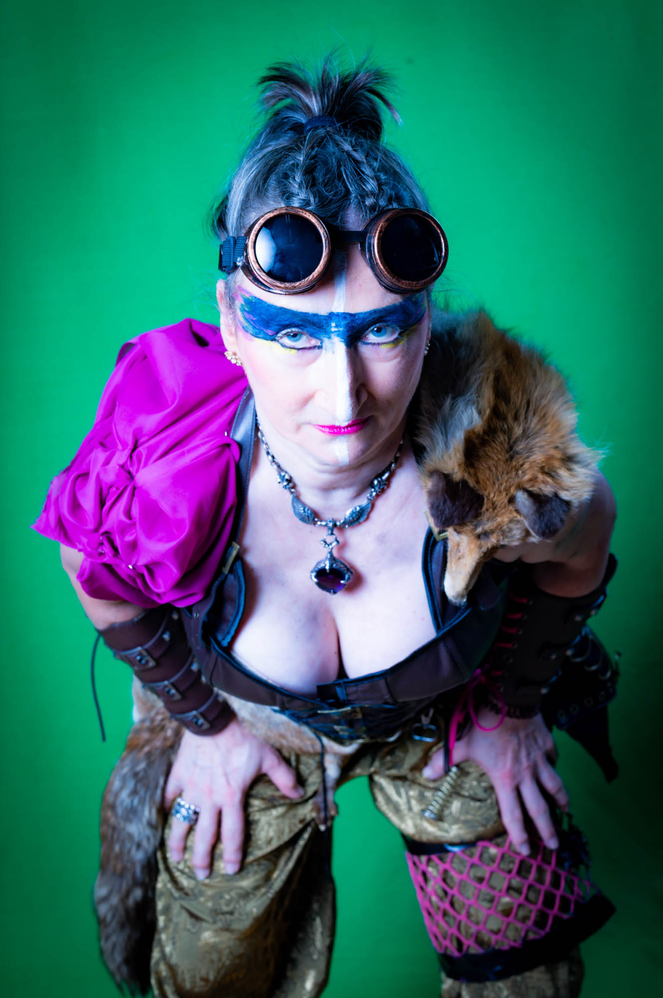 Steampunk fashion with colorful makeup, goggles, fur accessory, and vibrant attire on a green background.