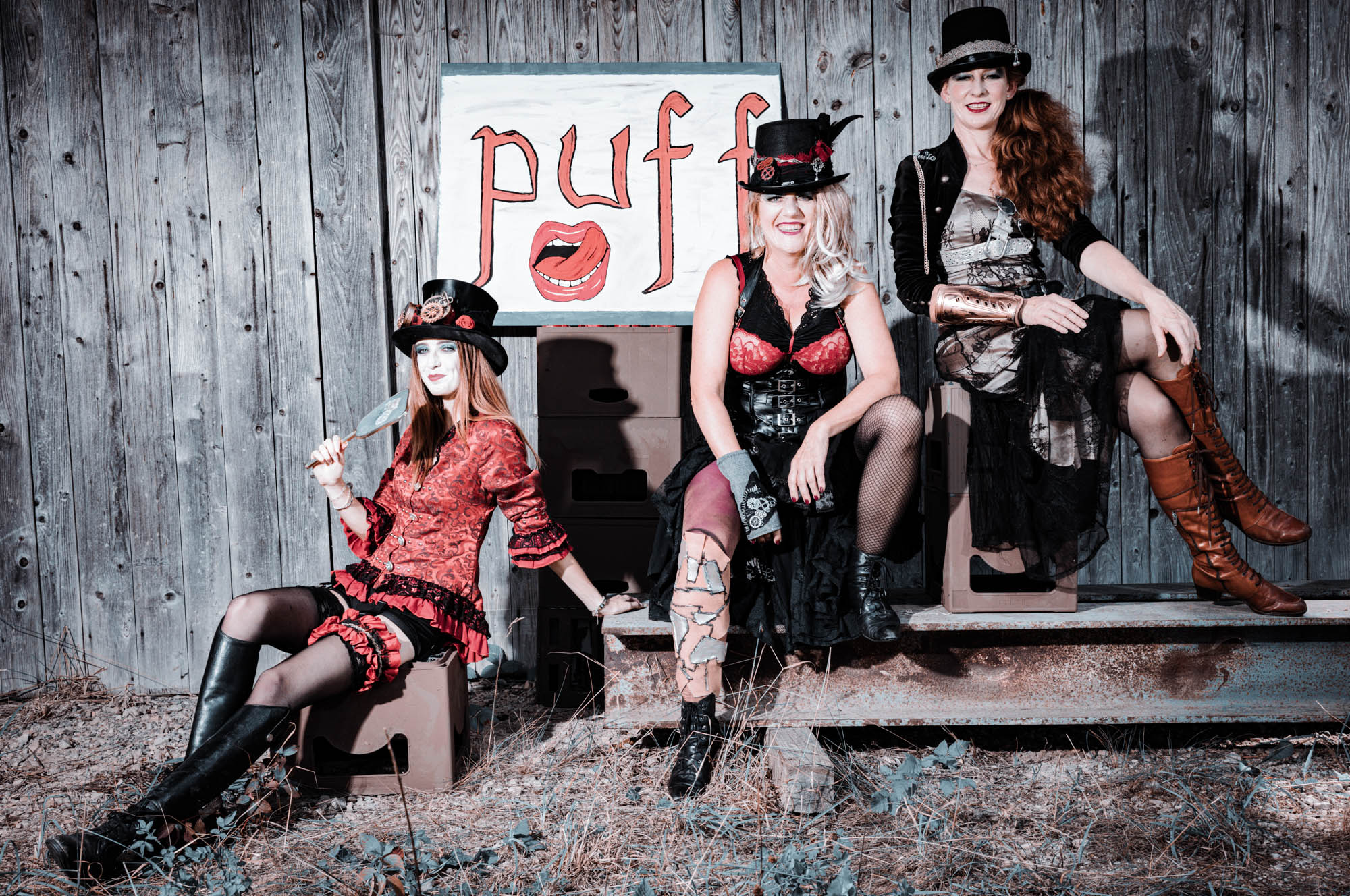 Three women in steampunk attire posing in front of a wooden wall with a puff sign.