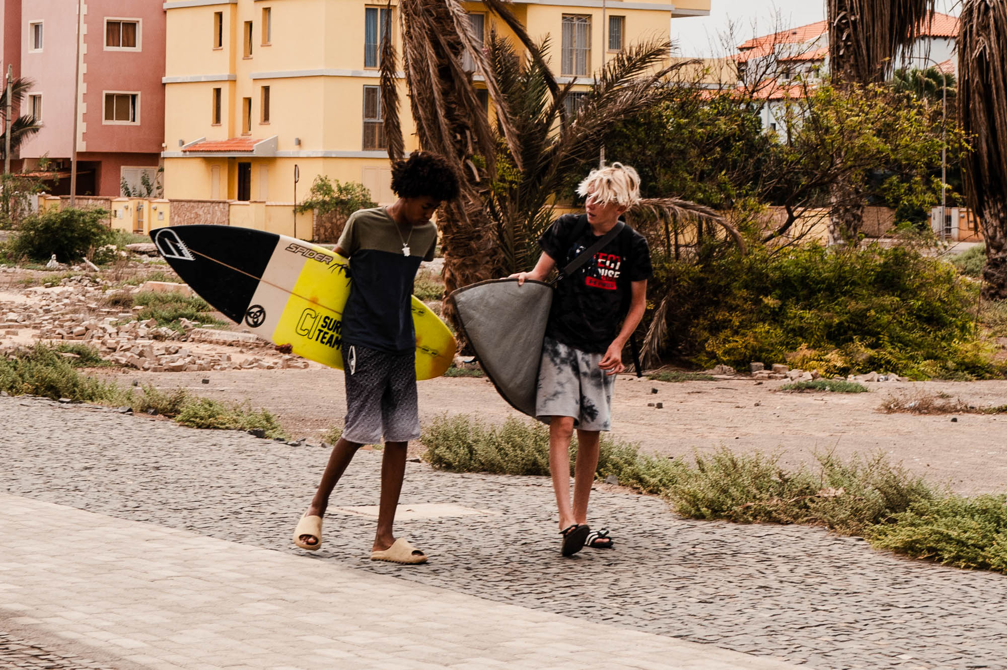 Two young surfers walking with boards near coastal buildings and palm trees, heading to the beach.