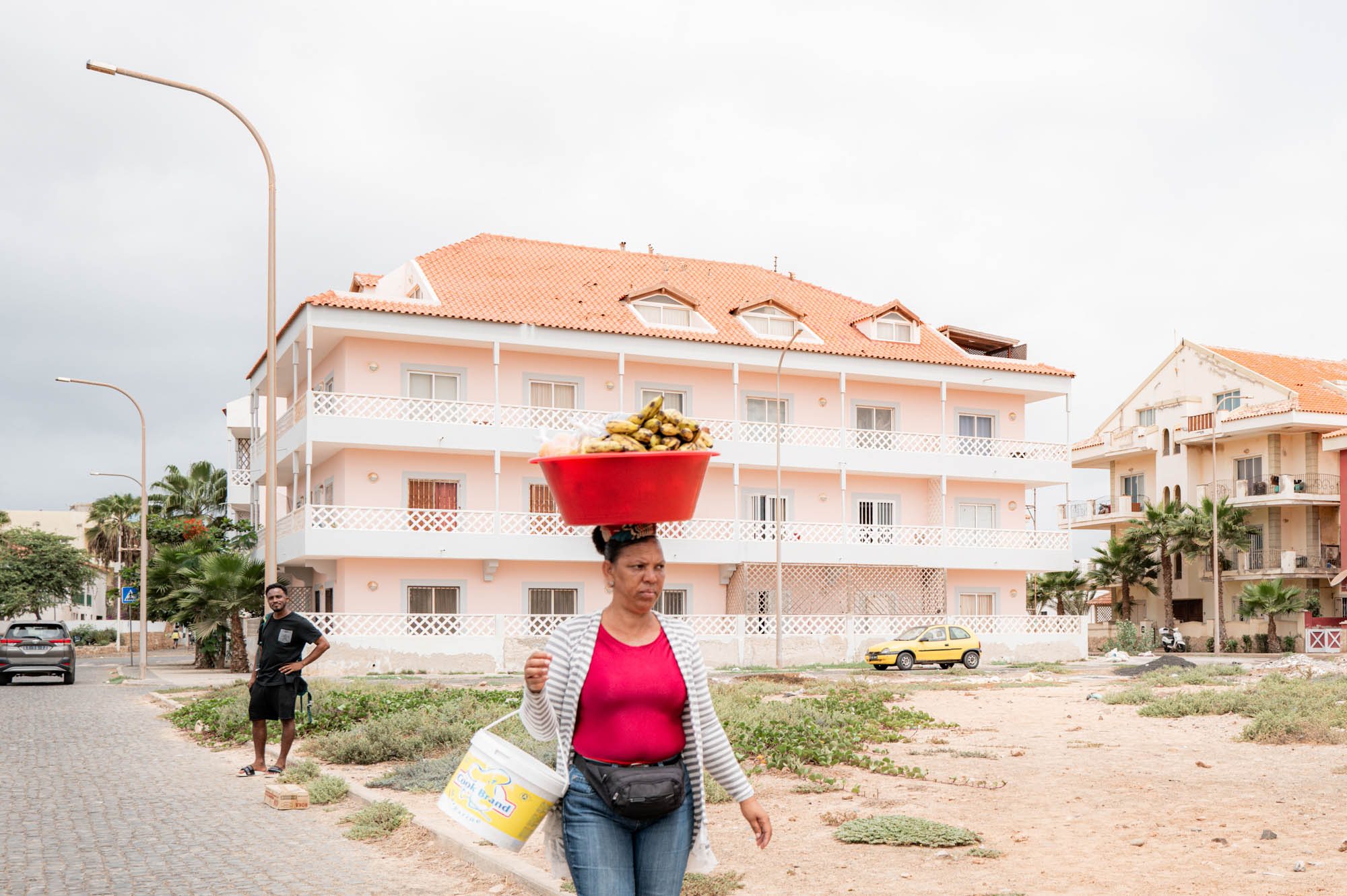 Woman balances a red basket on her head near pastel buildings, showcasing street life and local culture.