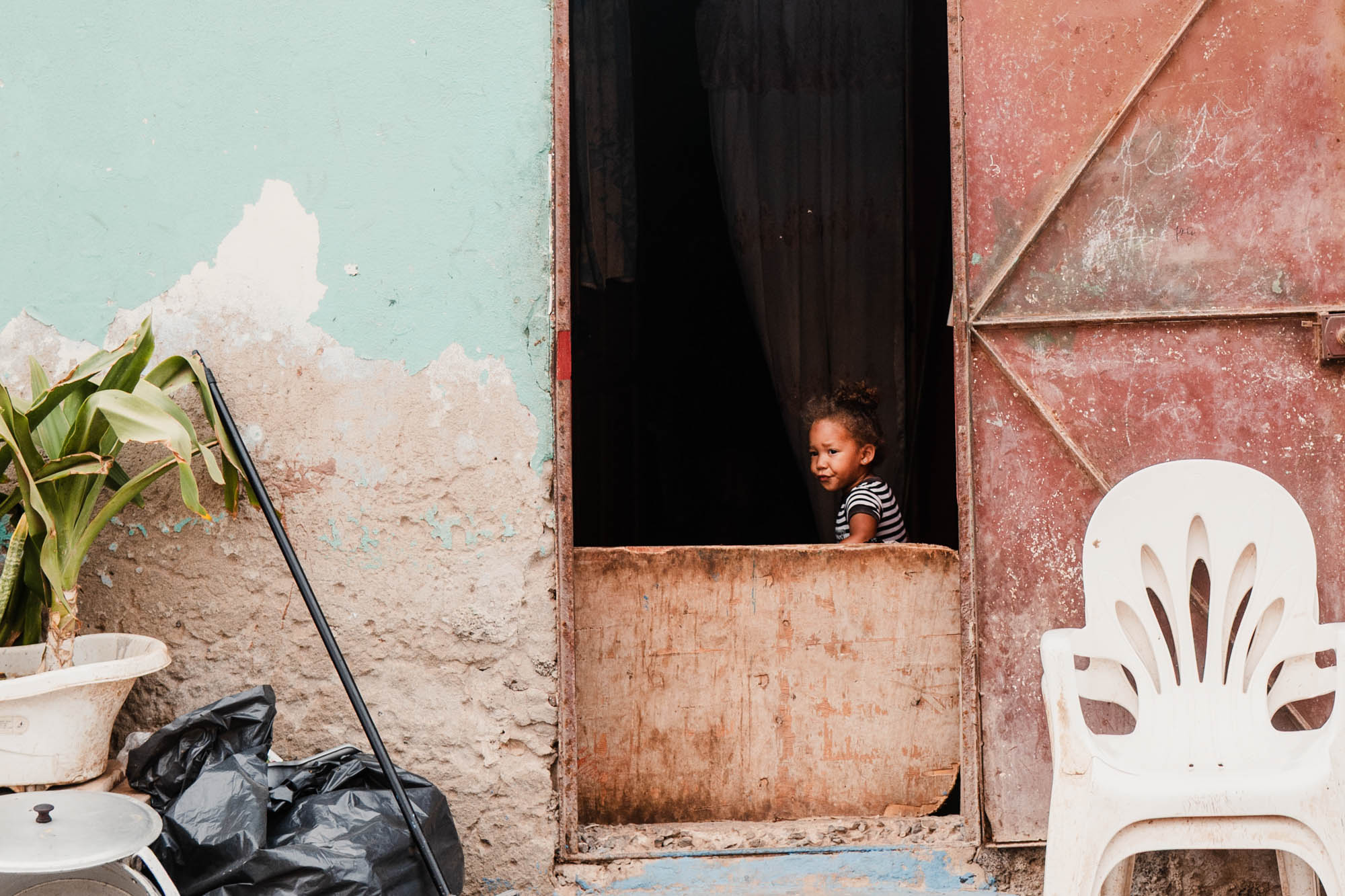Child peering out a rustic wooden door of a weathered building, with peeling paint and outdoor items nearby.