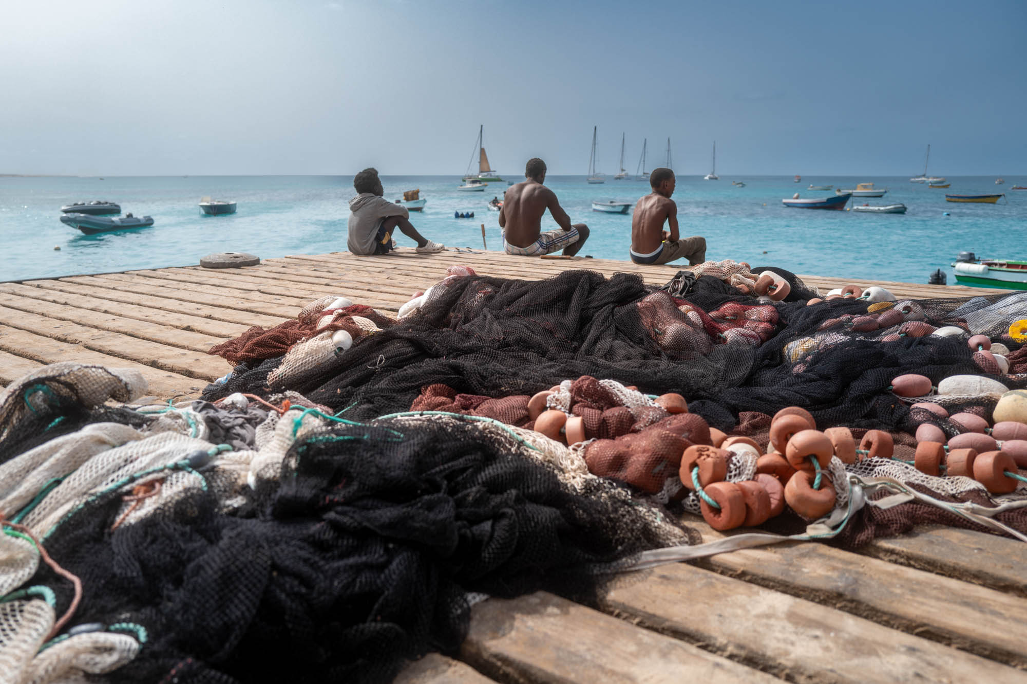 Fishing nets on a pier with kids sitting, looking at boats on a calm sea under a clear blue sky.