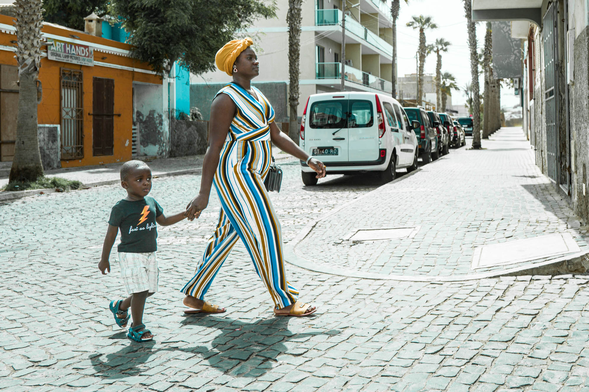 Mother and child walking on a sunlit cobblestone street, both dressed in colorful outfits.
