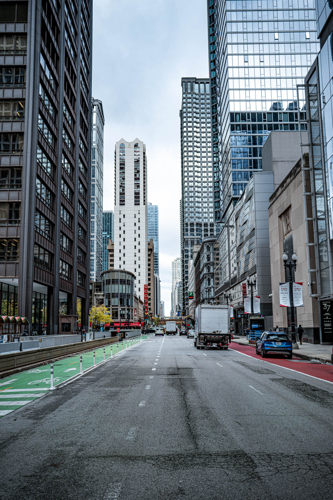 Downtown city street with tall skyscrapers, vehicles, and a bike lane, under a cloudy sky.