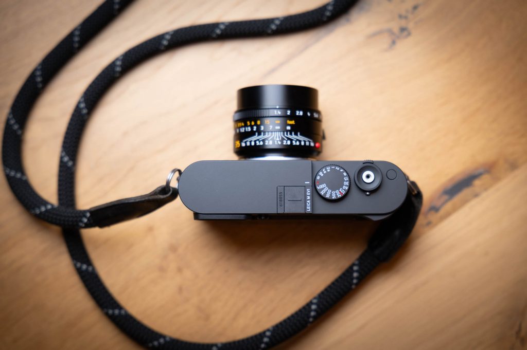 Compact black camera with a textured strap on a wooden surface, featuring a manual control dial and lens side view.