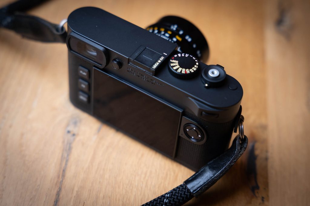 Black digital camera with a strap, on a wooden table, showcasing multifunction dials and a rear display.